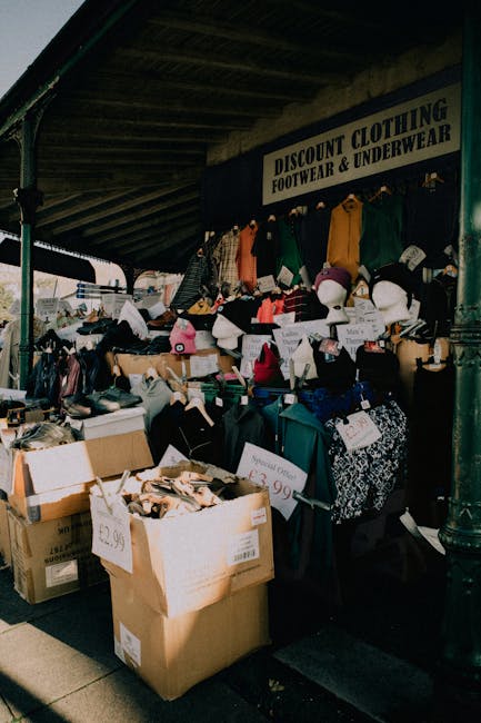 Outside a second-hand clothing stall with a sign advertising discount footwear and underwear, the shop displays an array of items including handbags, hats, and clothing on shelves and hangers. Several