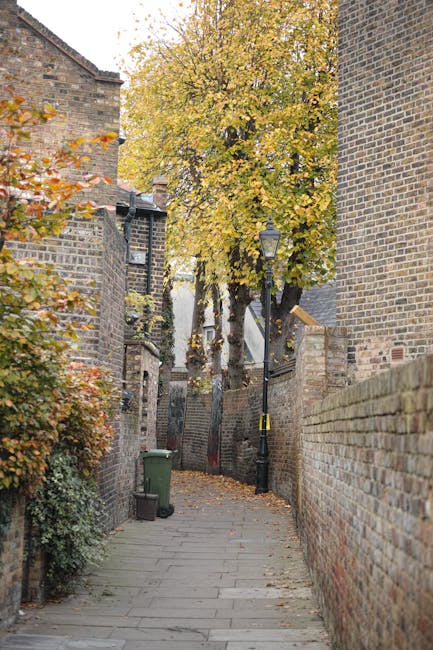 A narrow residential street paved with grey stone slabs, bordered by high brick walls on both sides, with some brickwork showing signs of age and weathering. A black street lamp with a decorative top stands on the right side near the middle of the image, and a green wheelie bin is positioned against the left wall. In the background, a large tree with yellow and orange autumn leaves overhangs the pathway, partially obscuring a white building at the end of the alley. The scene is lit by natural daylight, highlighting the textures of the brickwork and the vibrant foliage. This setting depicts a typical urban passageway suitable for house removals or furniture transport, with the street's confined space presenting logistical considerations during a home relocation, which [COMPANY_NAME], such as Wandsworth Movers, may assist with for moves within Wandsworth or similar areas. The context suggests a peaceful neighborhood ready for a moving or packing process.
